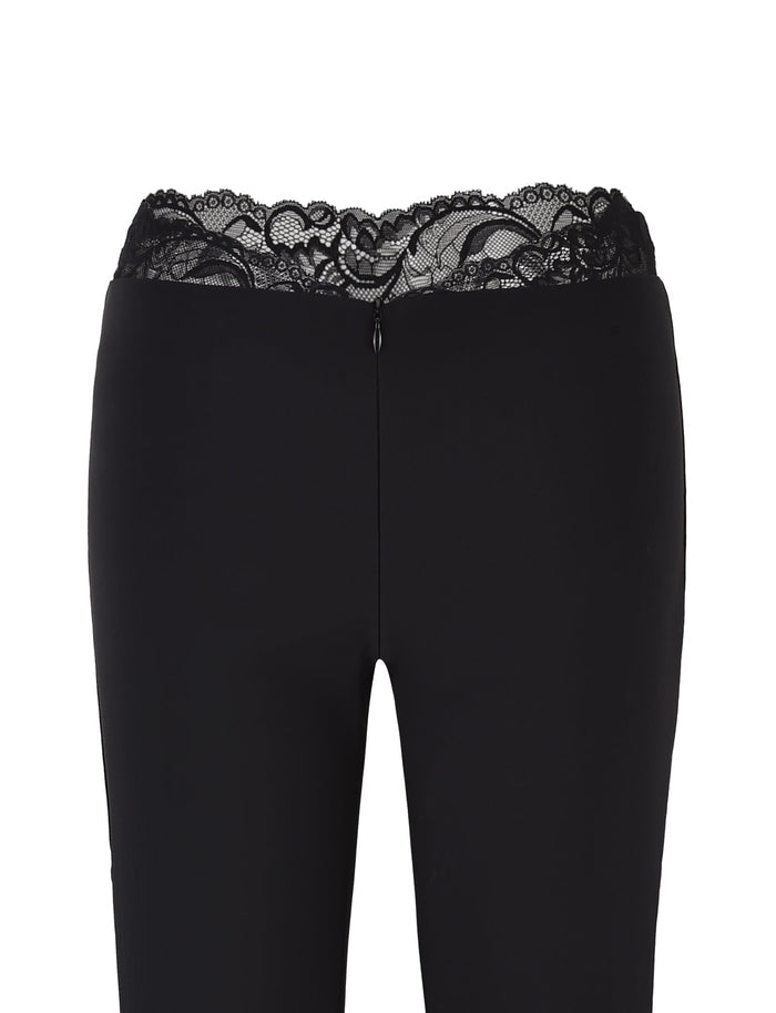 The NYOMI PANT - BLACK by TIGERMIST are black flared leg pants with a high waist and lace trim at the top, made from high stretch fabric and featuring a rear zipper closure.