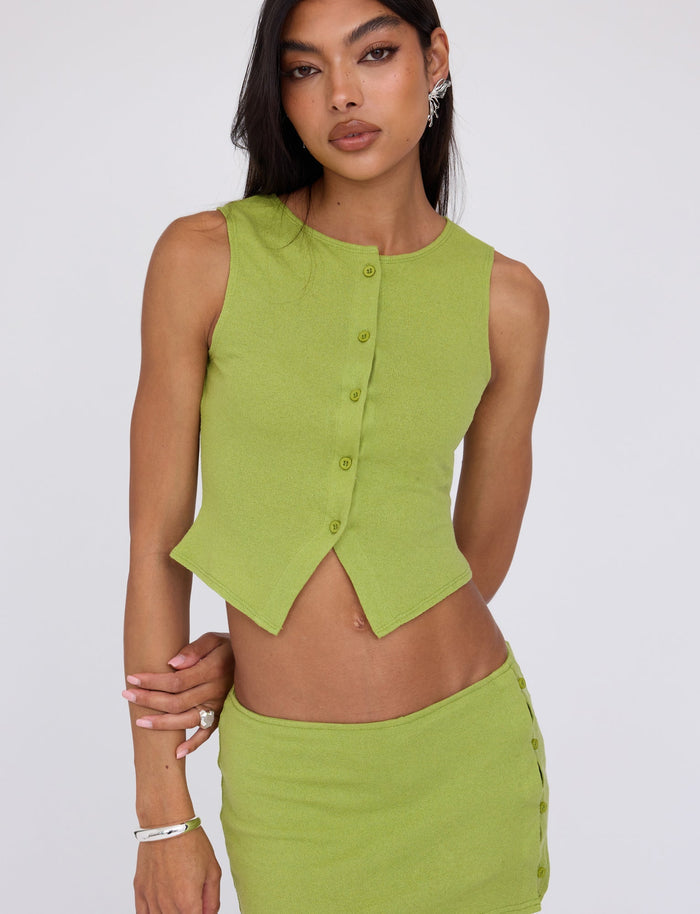 A woman with long hair is wearing the Matcha Green OAKLEE SKORT from TIGERMIST. She is donning a sleeveless, button-up top with a distinctive pointed hem paired with a matching green skirt featuring side buttons. She stands against a plain white background, looking into the camera.