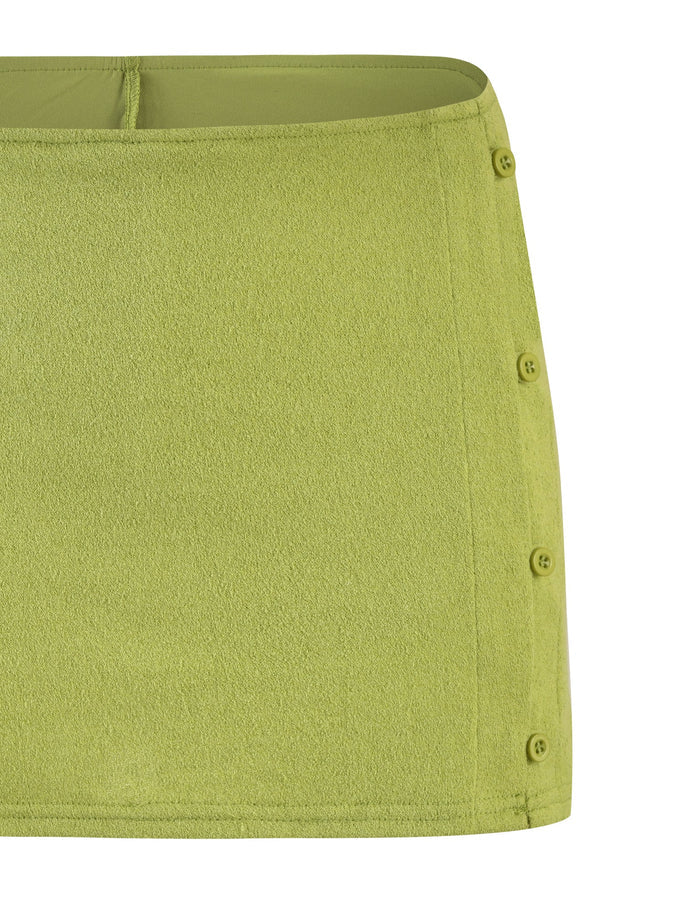 A close-up of the OAKLEE SKORT in Matcha Green from TIGERMIST shows its textured fabric and four vertically aligned buttons on the right side. The skort features a straight hemline and a minimalist design.
