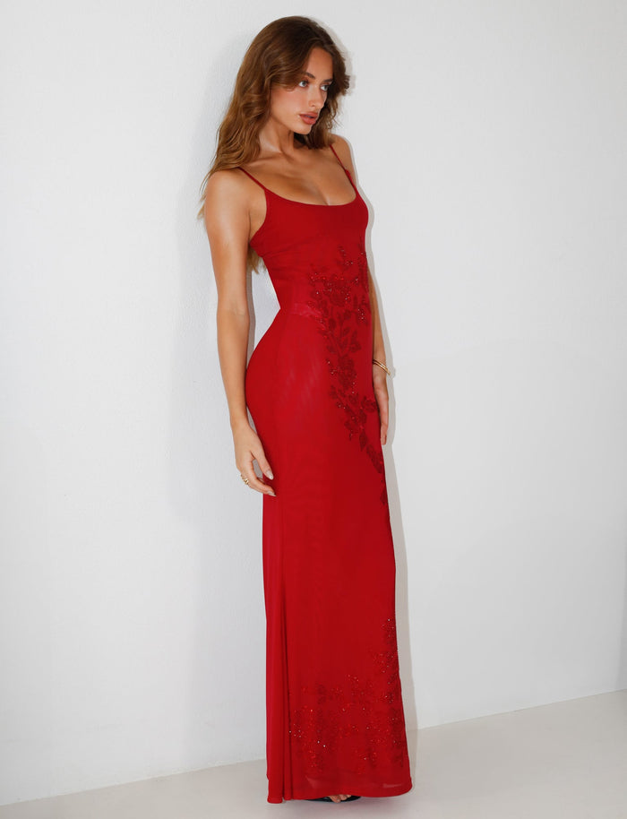 A woman in the OCTAVIA MAXI DRESS - CHERRY RED by TIGERMIST, a sophisticated long red dress with thin straps and delicate floral details, stands against a plain white background. The maxi-length piece beautifully accents her long, wavy hair as she poses with relaxed elegance.