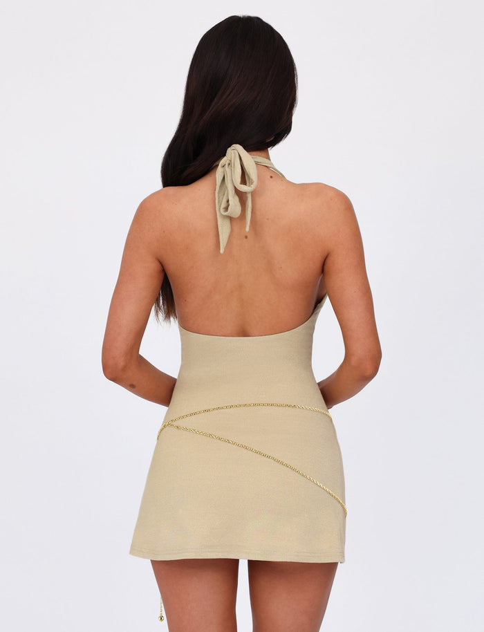 A woman with long dark hair is standing with her back to the camera, wearing the OPELI DRESS - GOLD from TIGERMIST. The short, light beige halter neck dress is backless and features thin gold chain details draping across her back and hips. The background is plain white.