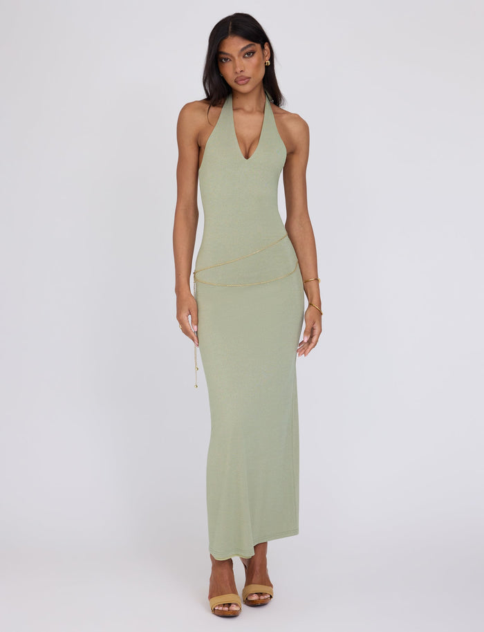 A woman stands against a plain background wearing a light green TIGERMIST Opeli Maxi Dress in Green Sparkle. She has long, straight hair and is accessorized with gold earrings, a delicate gold chain belt at the waist, and gold high-heeled sandals. Her expression is neutral, and her arms rest by her sides.