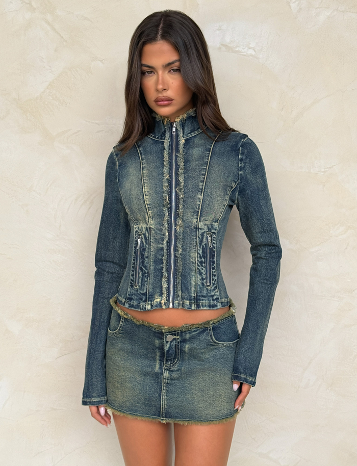 A woman poses against a beige textured wall, wearing the TIGERMIST OPHELIE JACKET in denim blue with frayed edges, paired with a matching short denim skirt.