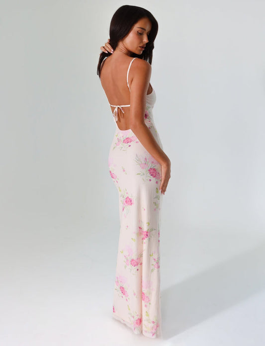 A woman stands against a plain background wearing the OSIRIS Dress by TIGERMIST. This long, open-back dress showcases a deep V neckline and features a watercolor floral print with pink and purple flowers on a white base. She is slightly turned to the side, with her arms by her sides, looking downward.