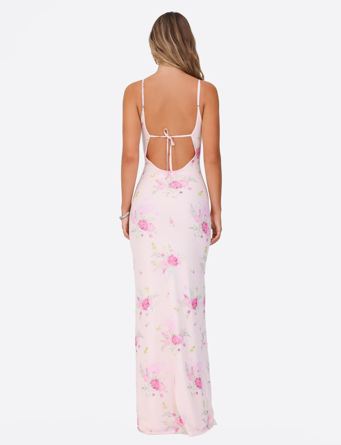 A woman with long hair is seen from the back wearing the TIGERMIST OSIRIS DRESS in a light pink, watercolor floral print. The dress, made of mesh fabrication, showcases a sleeveless design with an open back tied with a bow and features a flowing maxi length. She stands against a plain white background.