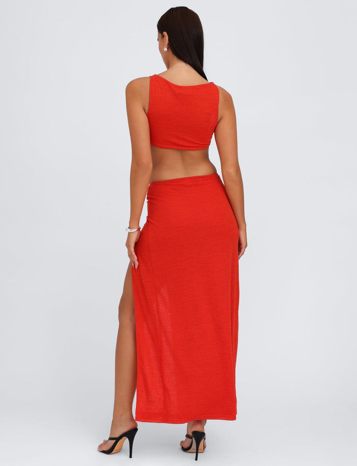 A woman with long dark hair, wearing a sleeveless red crop top and the TIGERMIST PAXTON SKIRT - RED : OMBRE with a high split, stands with her back to the camera. She is also wearing black high-heeled sandals and a bracelet on her right wrist.