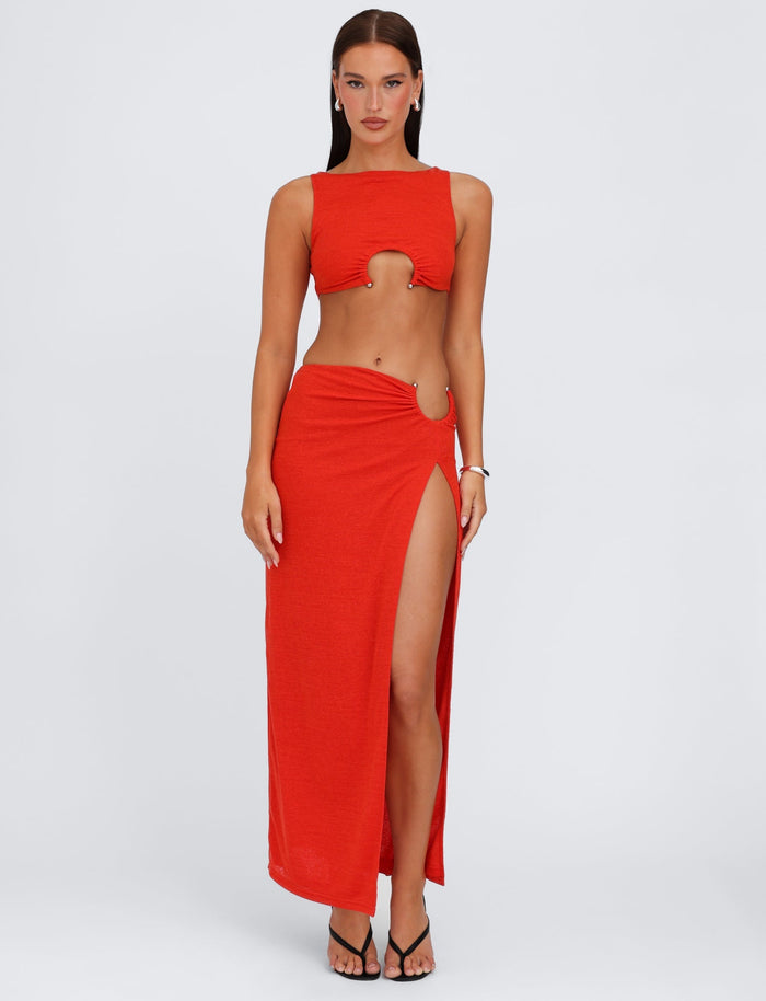 A woman stands against a plain white background wearing a vibrant red two-piece outfit from TIGERMIST. The ensemble consists of the PAXTON SKIRT - RED : OMBRE, featuring a crop top with a keyhole cutout and a high split, low-rise waist maxi skirt. She complements the look with black high-heeled sandals and a bracelet. Her hair is straight and parted in the middle.