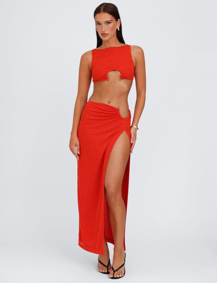 A woman poses confidently in a matching red crop top and the TIGERMIST PAXTON SKIRT - RED: OMBRE, with her left hand on her hip. The crop top features a circular cutout on the front, and she is wearing black high-heeled sandals. The background is plain white.