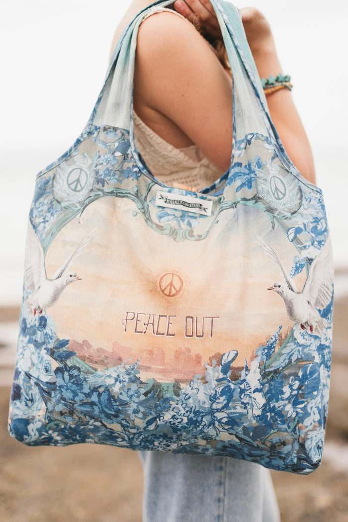 Model wearing our Peace Out tote bag featuring 1800's perfumeur sign with gentle tones of blue & aqua, landscapes, clouds, dove, and peace symbols -Market of Stars.