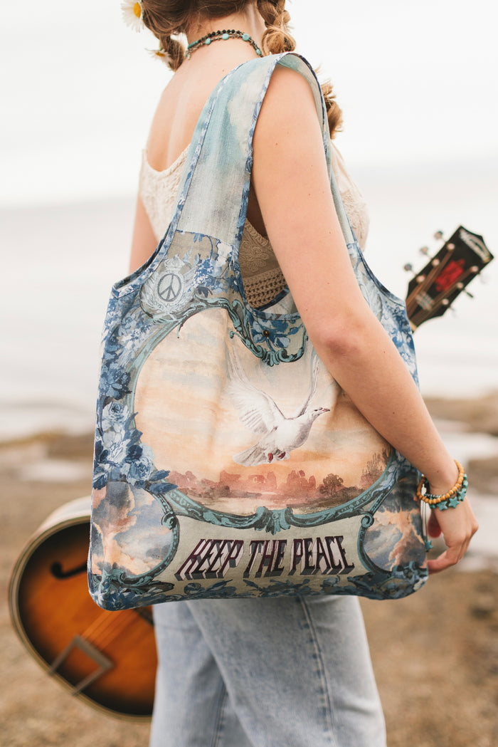 Model wearing our Peace Out tote bag featuring 1800's perfumeur sign with gentle tones of blue & aqua, landscapes, clouds, dove, and peace symbols -Market of Stars.