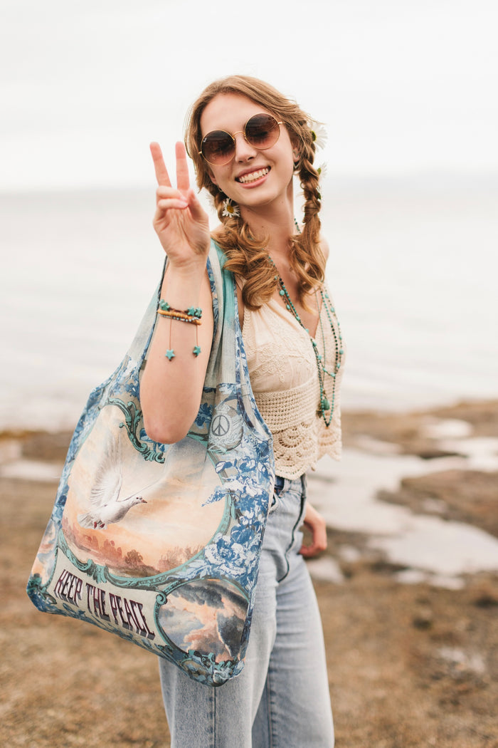Model wearing our Peace Out tote bag featuring 1800's perfumeur sign with gentle tones of blue & aqua, landscapes, clouds, dove, and peace symbols -Market of Stars.