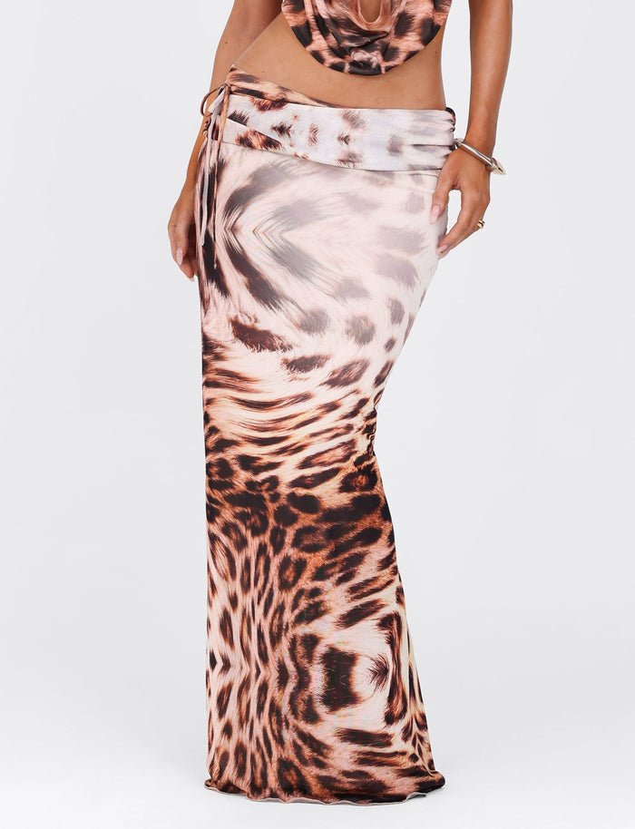 A person wearing the TIGERMIST PEARL MAXI SKIRT - NEUTRAL : LEOPARD : LEOPARD HAZE stands against a plain background. The leopard colorway includes brown, beige, and black patterns. The partially visible top complements the skirt's design, and the person's arm is adorned with several bracelets.