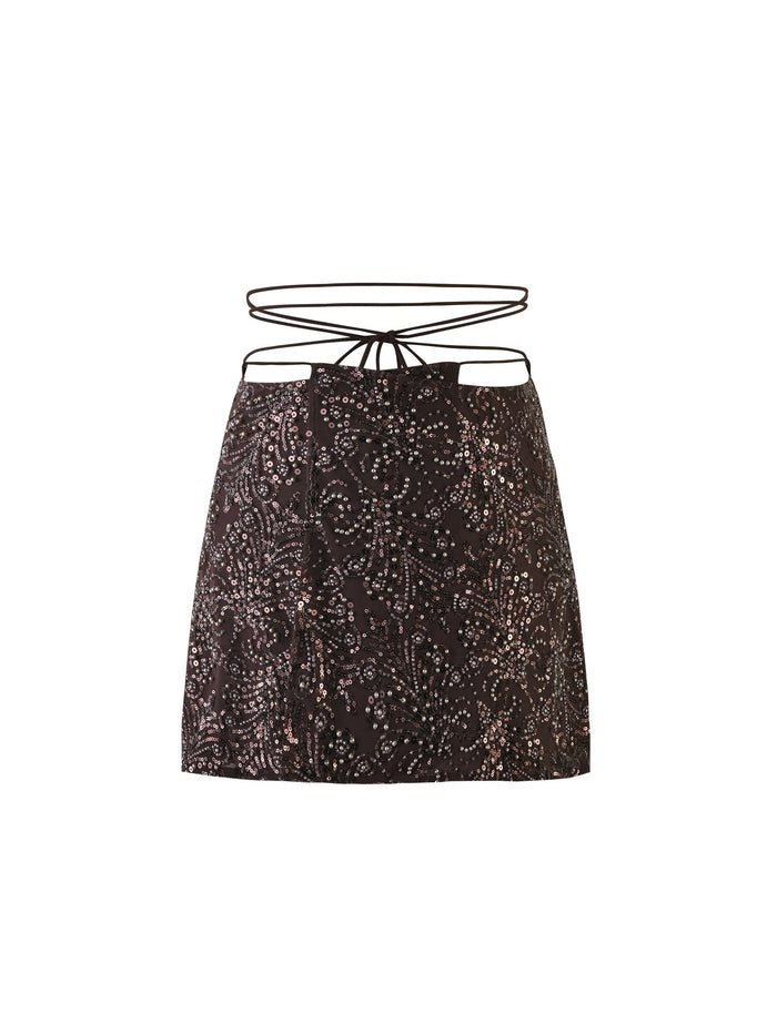 The RAINA SKIRT in chocolate brown from TIGERMIST boasts a detailed white and gray sequin pattern. It is stylishly adorned with thin black string ties at the waist, adding an intricate design element above the top hem of the skirt.