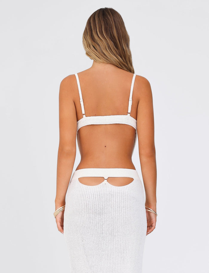 A woman with long hair is elegantly dressed in the TIGERMIST Ramsey Top - White, a chic knit two-piece featuring a crochet top with adjustable straps. The outfit includes a stylish skirt with twin cutouts at the waist, against a plain background, showcasing effortless elegance.
