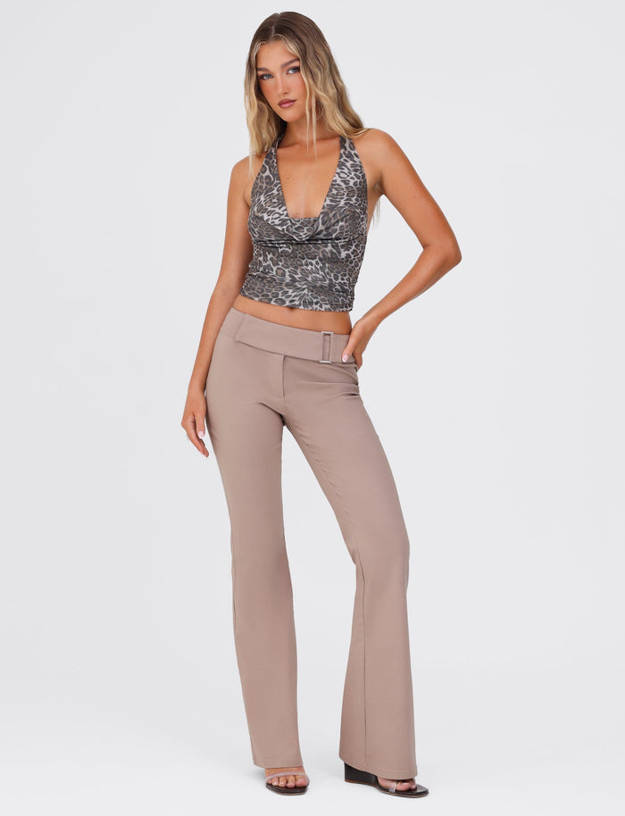 A person with long hair stands confidently, wearing the RILYNN TOP - NEUTRAL : LEOPARD from TIGERMIST, a sleeveless deep V-neck leopard-print top, paired with high-waisted beige flared pants featuring a belt. The high-stretch pants accentuate their figure perfectly. They complete the look with black open-toe heels. The backdrop is a plain white studio setting.