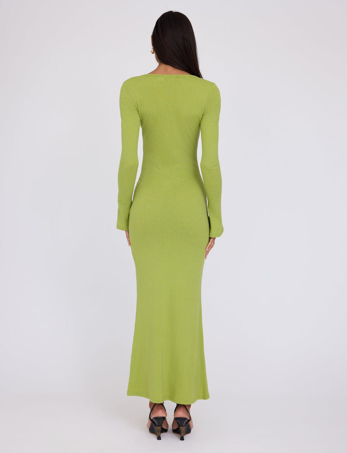 A person with long dark hair stands facing away, wearing the floor-length, fitted ROMIE DRESS in Matcha Green from TIGERMIST. The individual is also wearing black high-heeled shoes. The background is plain white, drawing attention to the clothes.