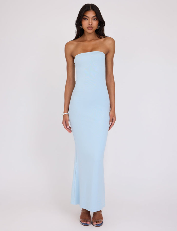 A woman stands against a plain white background wearing the TIGERMIST ROMINA DRESS in blue. She has long, dark hair and is sporting open-toed high heels. Her arms hang relaxed at her sides, and she maintains a neutral expression on her face.