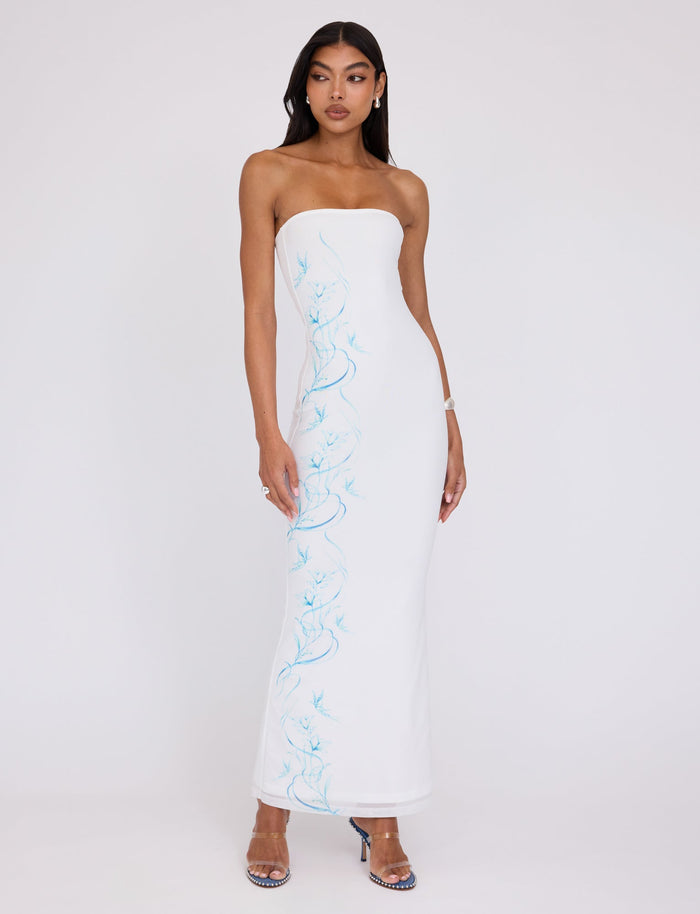 A woman stands against a pale background, modeling the ROMINA MESH DRESS - WHITE by TIGERMIST. The long, strapless dress is adorned with delicate blue floral patterns along the side. She accessorizes with blue open-toe heels and small earrings, wearing her hair down and displaying a serene expression on her face.