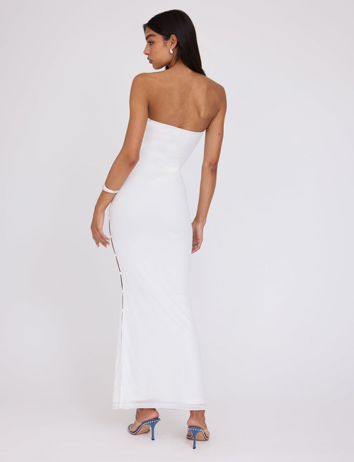 A woman with long, dark hair stands with her back to the camera, wearing the ROMINA MESH DRESS – WHITE from TIGERMIST. This strapless, form-fitting gown reaches the floor and features a side slit that reveals her legs as she poses. She is also wearing blue high-heeled shoes and bracelets.