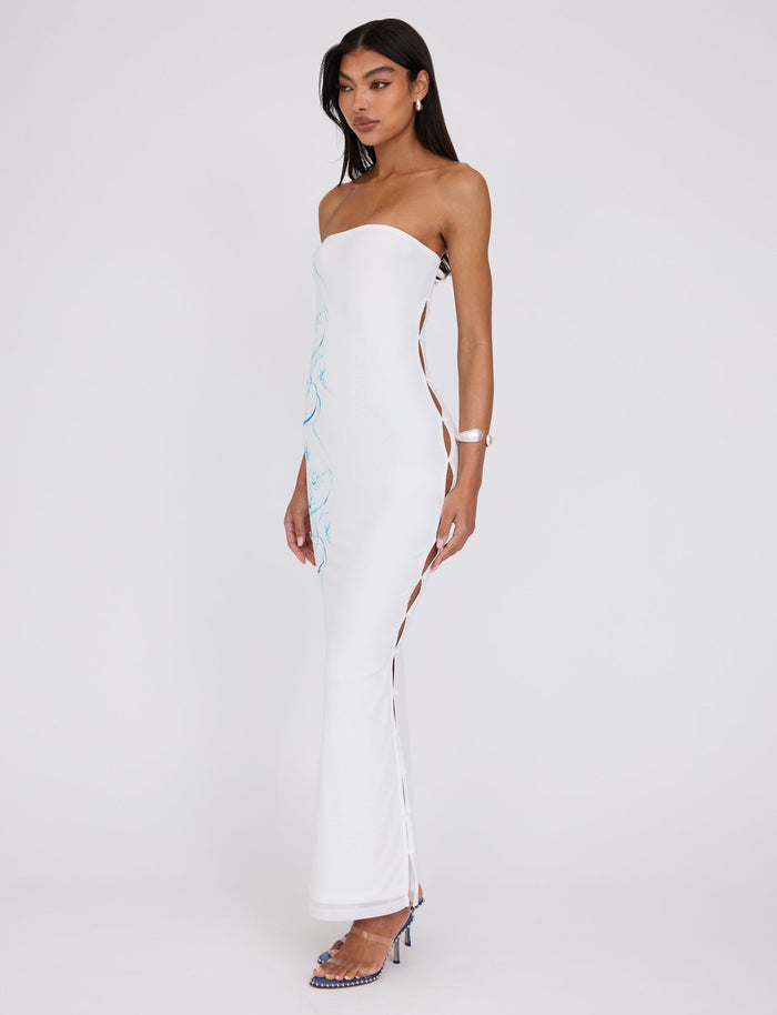 A woman stands against a plain background wearing the ROMINA MESH DRESS in white by TIGERMIST, which features side cut-outs and long, lace-up detailing. She has long dark hair and is wearing high-heeled blue sandals, complemented by a bracelet on her right wrist.