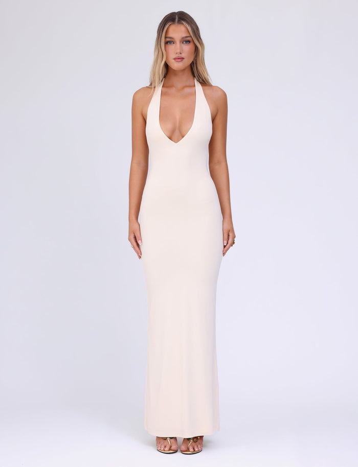 A woman stands against a plain background wearing the form-fitting ROZLYNN DRESS by TIGERMIST in a butter-yellow shade. The gown features halter straps and a deep V neckline, elegantly complementing her long, wavy hair. She pairs it with high-heeled sandals, facing the camera with a neutral expression.