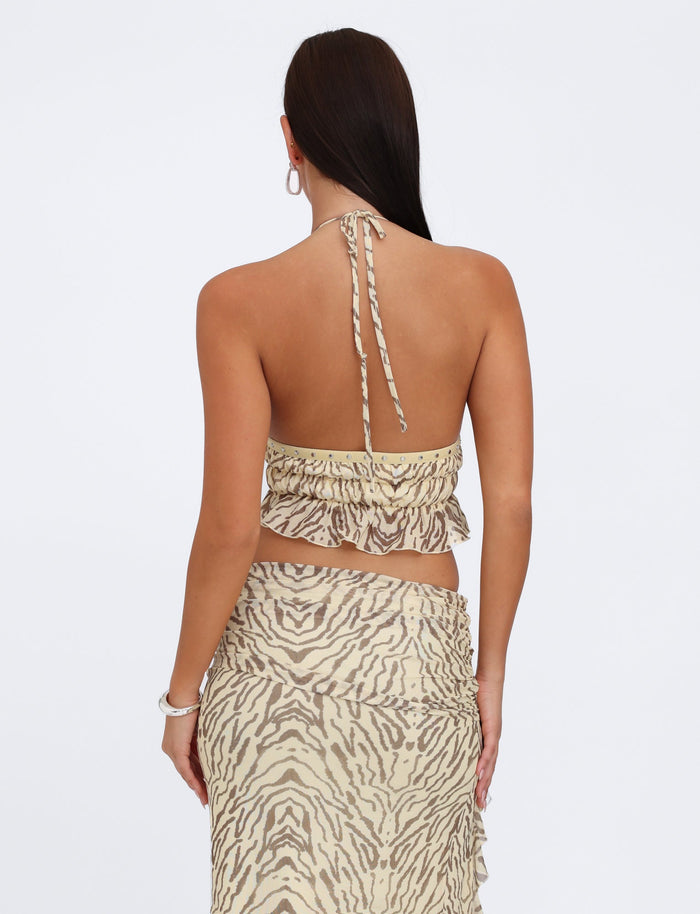 A woman with long dark hair is shown from the back, wearing the RUMER TOP - YELLOW : ZEBRA and a matching skirt from TIGERMIST. The top, featuring adjustable bust panels and ruffled detailing, is tied around her neck, while the skirt wraps around her waist with a ruffled hem. She accessorizes with hoop earrings and a bracelet.