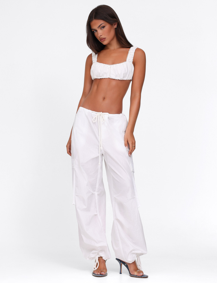 A woman poses against a plain white background wearing the SABRA TOP - WHITE by TIGERMIST, paired with loose white drawstring pants and embellished high-heeled shoes.