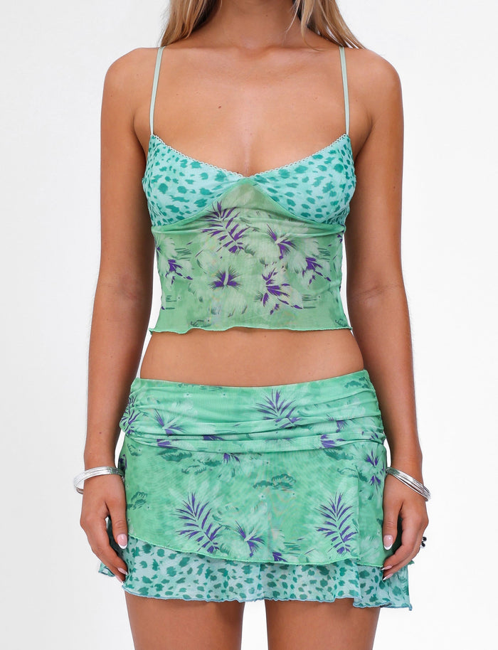 A person is wearing the SAIGE SKIRT - GREEN : FLORAL : ANIMAL two-piece outfit from TIGERMIST, featuring a sleeveless, fitted top and a matching short skirt. The high stretch fabric showcases beautiful floral and animal print designs. They are accessorized with some bracelets against a plain white background.
