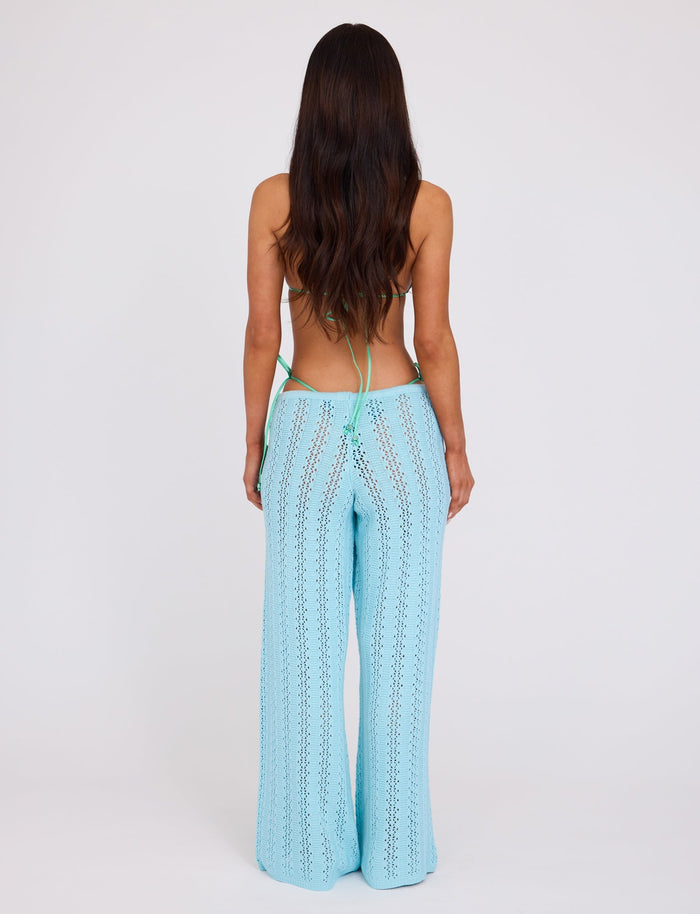 A woman with long, wavy brown hair stands facing away, wearing light blue SANDOR PANT - BLUE : BABY BLUE wide-leg crochet pants and a green strappy bikini top from TIGERMIST. The background is plain white.