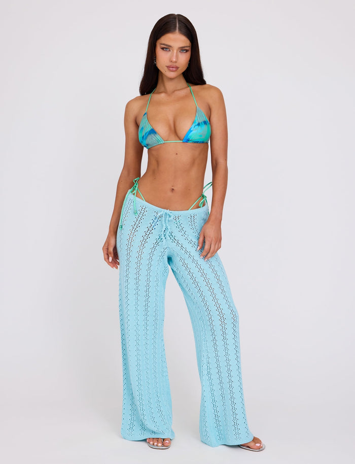A woman stands confidently against a plain background, wearing a TIGERMIST blue and green tie-dye bikini top paired with the TIGERMIST SANDOR PANT in BABY BLUE. She has long dark hair, minimal makeup, and poses with her hands by her sides, projecting a cool and stylish vibe.