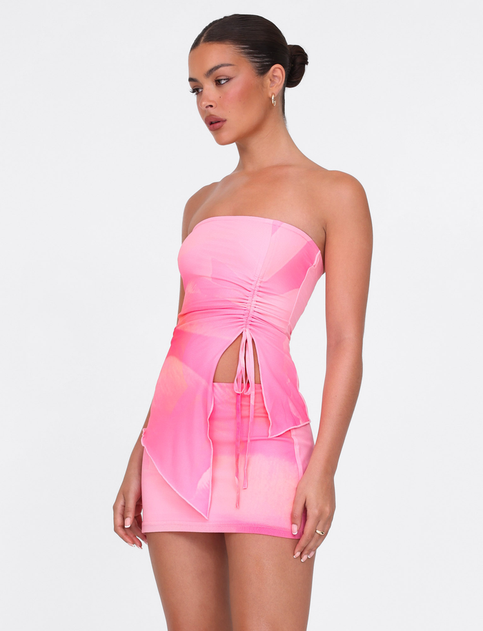A woman wears the TIGERMIST SARIAH MINI SKIRT in Macro Floral Pink Blossom—a strapless, form-fitting mini with a ruched front, side cutout, layered skirt detail, and high-stretch fabric—against a white background.