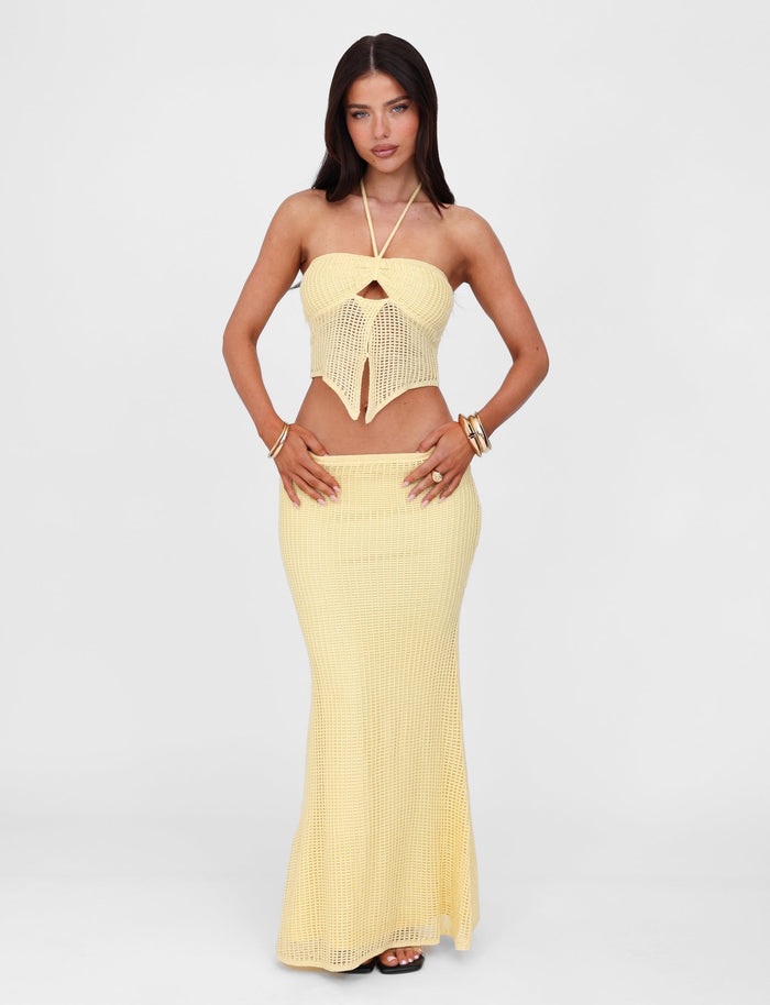 A woman stands against a plain background, wearing a matching knit outfit from TIGERMIST consisting of a yellow halter top and the SHAH SKIRT - YELLOW : LEMON. She accessorizes with several bracelets on her right wrist and strappy high heels. Her hair is straight and dark, and she has a neutral expression.