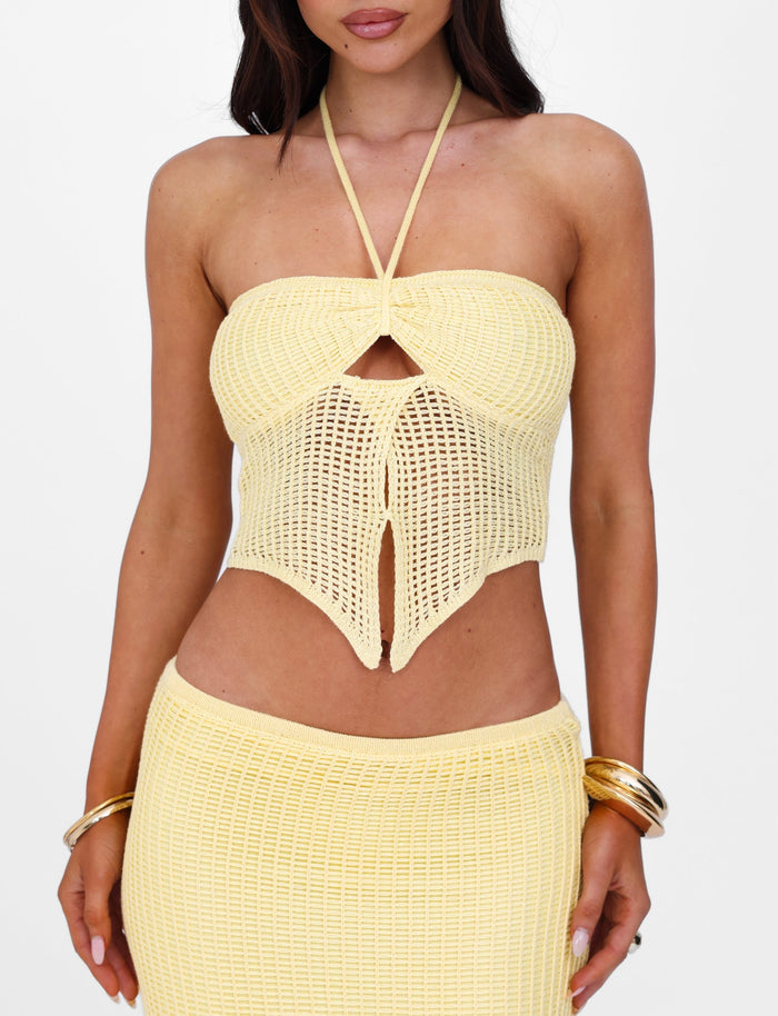 A woman is wearing the TIGERMIST SHAH TOP - YELLOW : LEMON, a matching yellow crochet halter top with a front keyhole cutout that can be styled in multiple ways. She pairs it with a hip-sitting skirt, has long dark hair, and accessorizes with gold bangle bracelets on both wrists against a plain white background.