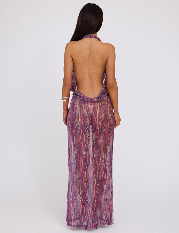 A person with long, dark hair is standing with their back to the camera, wearing the SONORA DRESS - MULTI : ROSE STRIPE from TIGERMIST, a backless, floor-length sleeveless dress featuring a purple, pink, and white swirling pattern. They are also wearing two silver bracelets on their left arm.