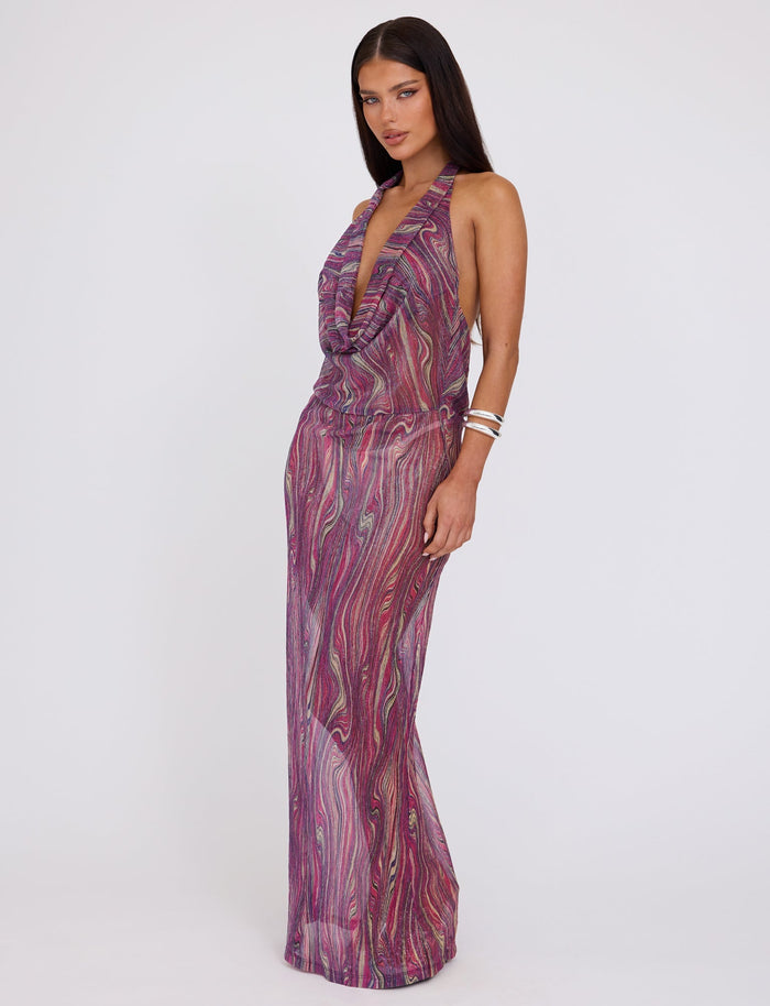 A woman stands against a plain white background, wearing the SONORA DRESS - MULTI : ROSE STRIPE from TIGERMIST. The sleeveless, floor-length dress showcases a colorful, swirling pattern in purple, pink, and white hues. It features a deep V-neckline and is complemented by several silver bracelets on her left wrist.