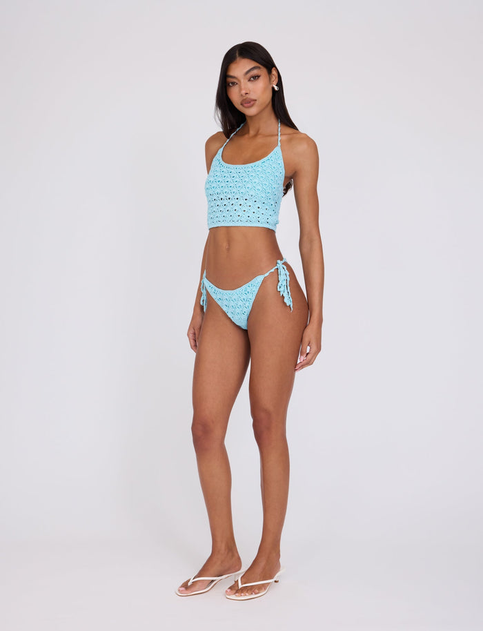 A woman is standing against a plain white background, wearing a light blue TIGERMIST crochet halter bikini top and matching SUNNIVA BIKINI BOTTOM - BLUE : BABY BLUE. She is looking at the camera with a neutral expression, with one leg slightly forward. She is also wearing flip-flops.