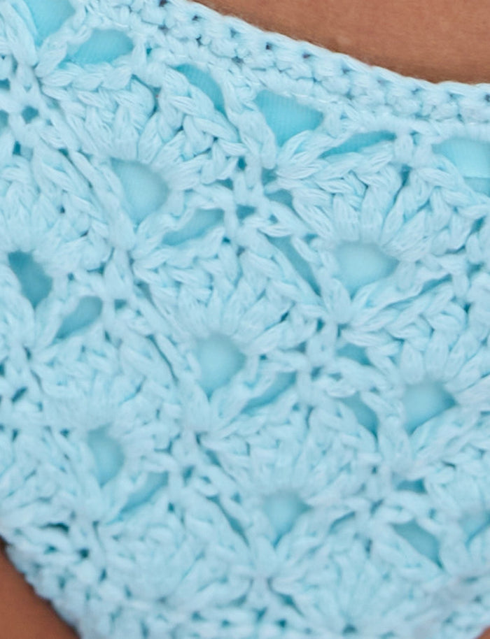 Close-up of the SUNNIVA BIKINI BOTTOM - BLUE: BABY BLUE by TIGERMIST, featuring a light blue crocheted fabric with intricate flower-like patterns. The yarn is woven in an airy, lacy style, highlighting the detailed craftsmanship. The background is out of focus and appears to be a person's skin.