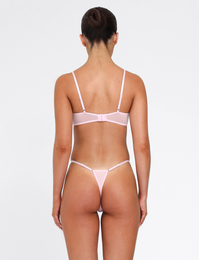 A woman with her hair in a bun stands with her back to the camera, wearing the TIGERMIST TALIYAH SET - PINK: BABY PINK—an elegant bra and thong in delicate embroidered mesh, set against a plain white background.