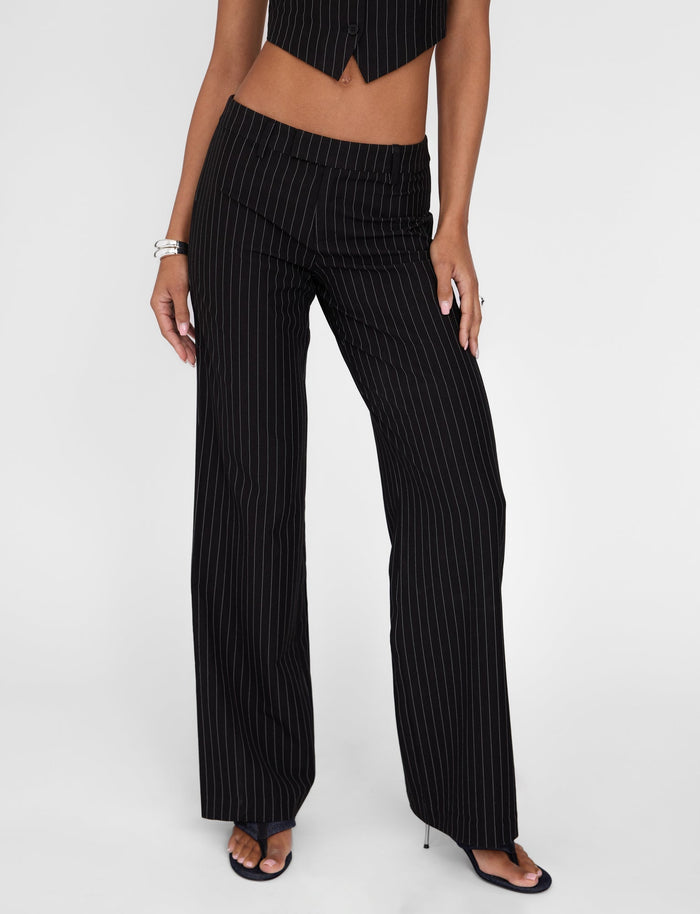 A person is seen wearing the TIGERMIST TAMARA PANT in black pinstripe with wide-leg design and a mid-rise waist, paired with a matching cropped vest. The outfit is completed with black open-toe heels and accessorized with a silver bracelet. The image captures only the lower half of the body against a plain white background.