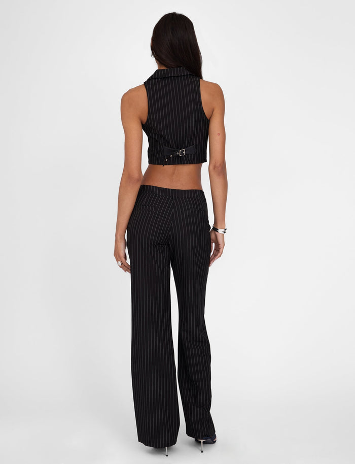 A woman stands facing away from the camera, showcasing TIGERMIST's black pinstripe sleeveless crop top paired with the TAMARA PANT - BLACK : PINSTRIPE. Her outfit includes a belt, and the Tamara Pant features a mid-rise waist and wide legs. She has long, dark hair and her hands are resting at her sides.