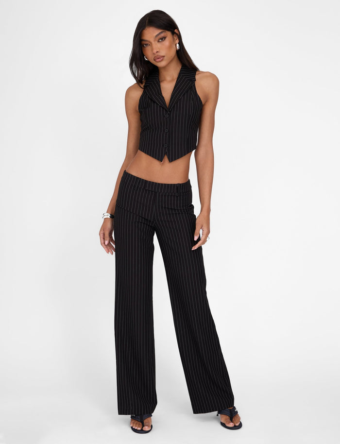 A woman stands against a plain white background, confidently posing in the TIGERMIST TAMARA VEST - BLACK : PINSTRIPE, a tailored fit ensemble featuring a cropped vest and high-waisted, wide-leg pants. She accessorizes with black heels, bracelets, and earrings. Her mid-length hair is down.