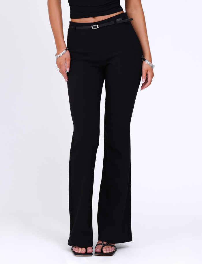 A person is wearing a black sleeveless top and the TIGERMIST TERLA PANT in black, which features a mid-rise waist and a flared silhouette. They stand with their hands relaxed at their sides, displaying silver bracelets on their left wrist. The flared pants complement their black strappy heels as they pose against a white background.