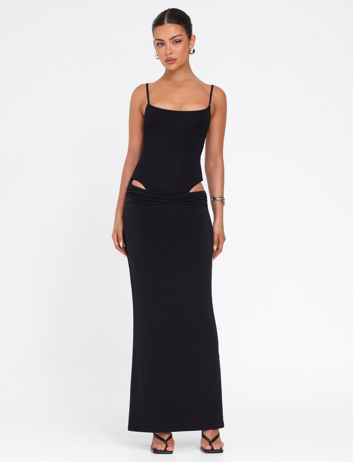 A woman poses against a white background in the UMA DRESS - BLACK by TIGERMIST, a fitted bodycon maxi dress with adjustable straps and hip cutouts. She completes her chic look with black heels, hoop earrings, and bangle bracelets.