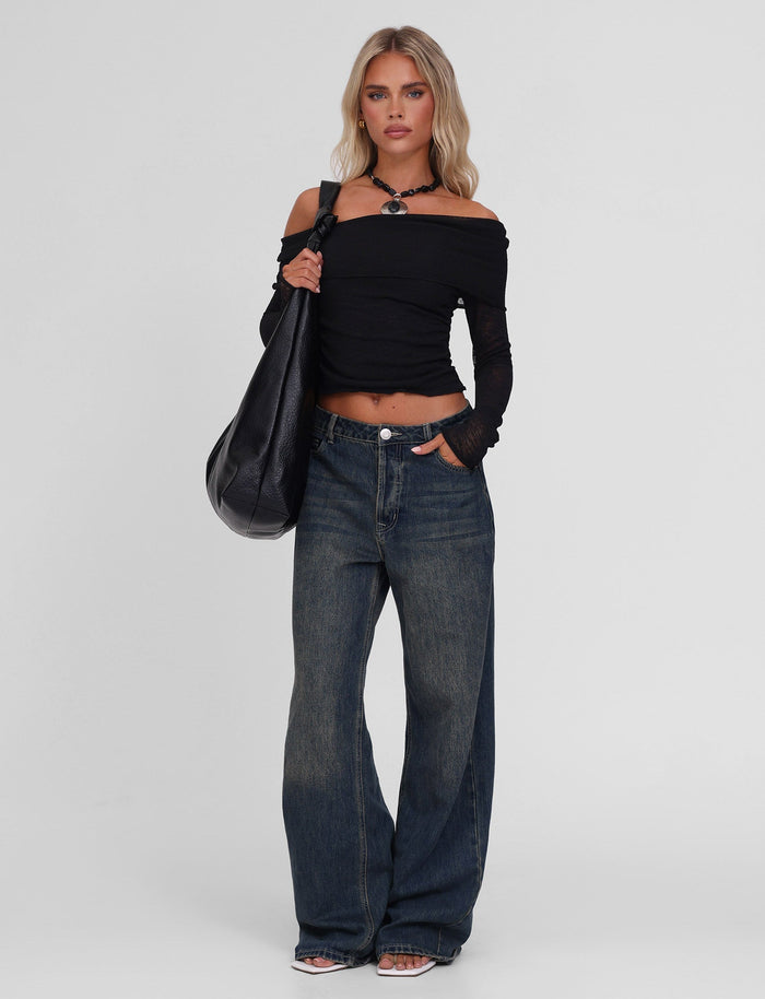 A woman stands against a plain background wearing the TIGERMIST VERA TOP in black, wide-leg blue jeans, clear-heeled shoes, a black choker, and carrying a large black shoulder bag.