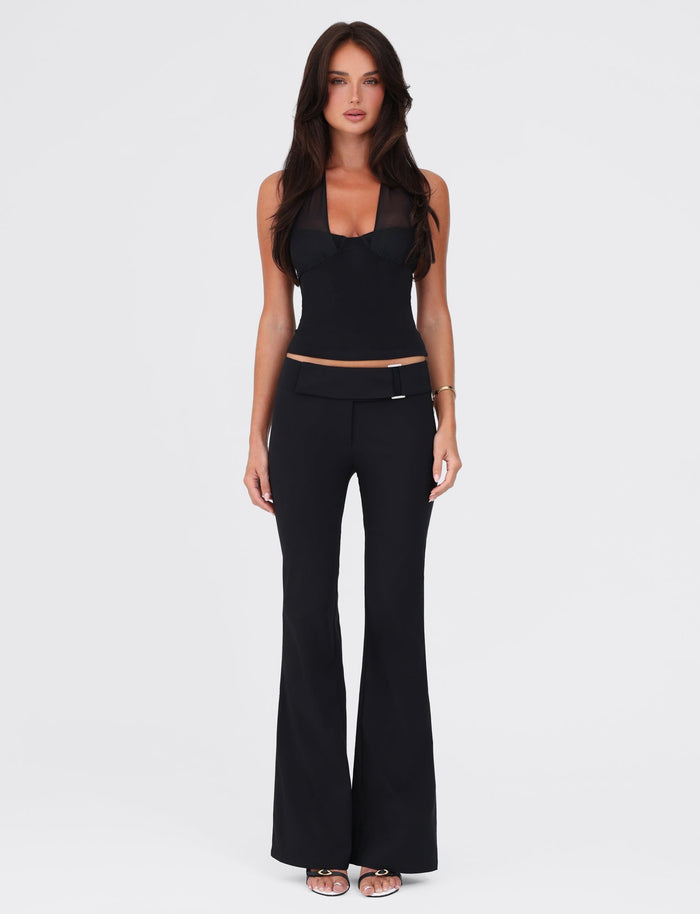 A woman with long, dark hair stands against a plain white background. She is wearing a black sleeveless top and the VESPER PANT - BLACK by TIGERMIST, featuring a fitted silhouette that accentuates her low-rise waist. With a neutral expression, her arms rest relaxed by her sides. She completes the outfit with black heeled sandals.