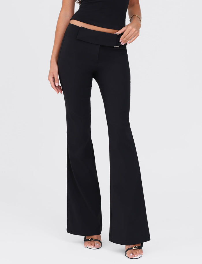 A person is wearing the VESPER PANT - BLACK by TIGERMIST, showcasing black flared pants with a fitted silhouette and a cropped black top. Their left hand rests by their side, while their right hand lightly touches their hip. The image captures only the lower half of the person, from the waist down to their feet, which are clad in open-toed shoes.