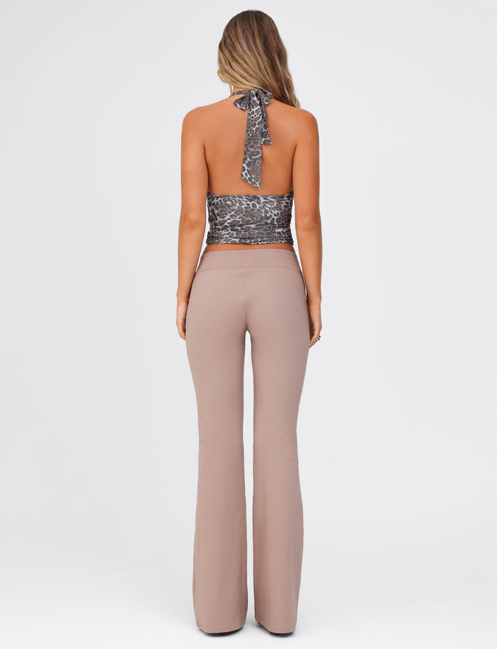 A woman is seen from the back, with long wavy hair. She is wearing a leopard print halter top that ties at the neck and TIGERMIST's VESPER PANT in brown and tan, which are high-waisted and flared. The background is plain and light grey.