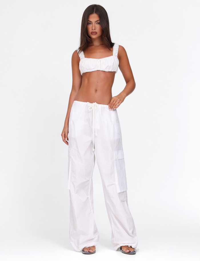 A woman with long, straight brown hair and a neutral expression stands against a white background wearing the TIGERMIST VIOLETTE PANT in white, styled with a white cropped top and flat sandals.