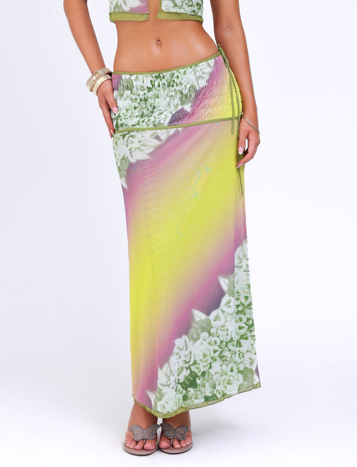 A woman is wearing a vibrant, colorful WAVERLY SKIRT by TIGERMIST, featuring a gradient of green with striped floral patterns and accentuating its low-rise waist. She is also wearing a matching top and sandals, with a bangle bracelet on her wrist against a plain white background.
