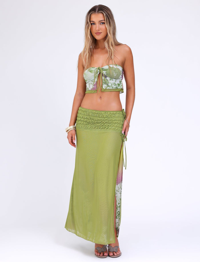 A woman with long blonde hair wears the TIGERMIST WAVERLY TOP - GREEN : FLORAL : STRIPE, a green floral tube top tied in the center and crafted from stretchy fabric. She pairs it with a matching high-waisted, ruched maxi skirt featuring a side slit. Accessorizing with white bracelets and sandals, she stands against a plain white background.